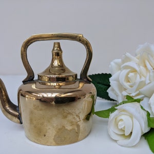 May include: A small, polished brass teapot with a rounded body, a curved handle, and a spout. The teapot is sitting on a white surface next to a bouquet of white roses.