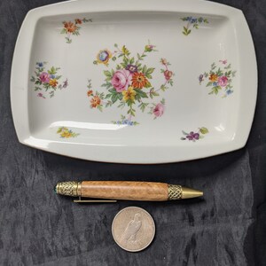 May include: A white rectangular serving dish with a floral pattern in shades of pink, yellow, and blue. The dish is on a black surface with a wooden pen and a silver coin.