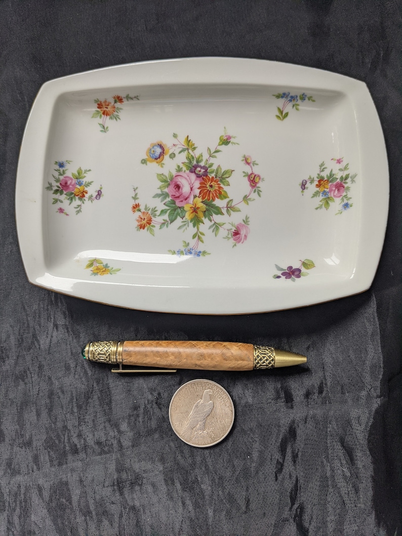 May include: A white rectangular serving dish with a floral pattern in shades of pink, yellow, and blue. The dish is on a black surface with a wooden pen and a silver coin.