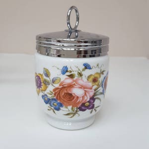 May include: A white ceramic egg cup with a floral design and a silver lid with a ring handle.