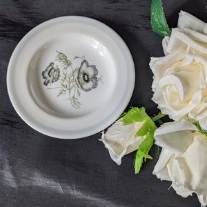 May include: A white ceramic saucer with a blue rim and a floral design featuring grey and green anemones. The saucer is sitting on a black surface next to white roses.