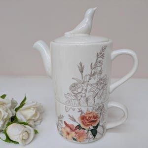 May include: A cream-coloured ceramic tea set consisting of a teapot stacked on a teacup. The teapot lid features a small bird figurine. Both pieces are decorated with detailed botanical illustrations of flowers in shades of orange, pink, and brown.
