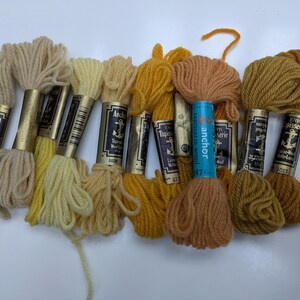 Pode incluir: Dez novelos de l&atilde; para tape&ccedil;aria Anchor em v&aacute;rios tons de amarelo e castanho. Os novelos s&atilde;o etiquetados com o nome da marca "Anchor" e o texto "Tapestry Wool".