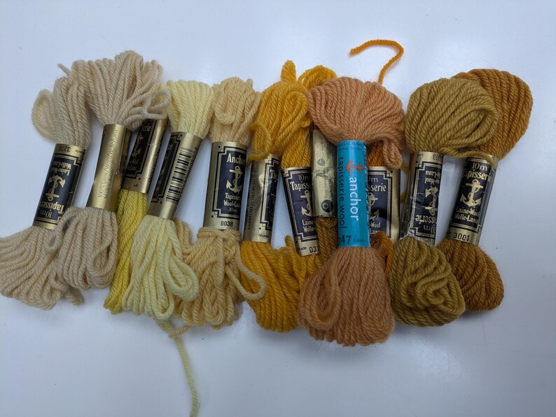 Pode incluir: Dez novelos de l&atilde; para tape&ccedil;aria Anchor em v&aacute;rios tons de amarelo e castanho. Os novelos s&atilde;o etiquetados com o nome da marca "Anchor" e o texto "Tapestry Wool".