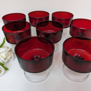 May include: Set of eight red glass dessert cups with clear glass bases. The cups are arranged in a circle on a white surface.