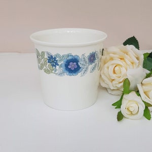 May include: A white ceramic planter with a blue and green floral design. The planter is sitting on a white surface with white roses.