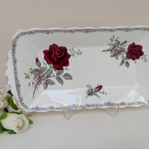 May include: A rectangular white serving platter with a scalloped edge and gold trim. It features burgundy roses, grey leaves, and small white flowers. The border is decorated with a repeating floral pattern.
