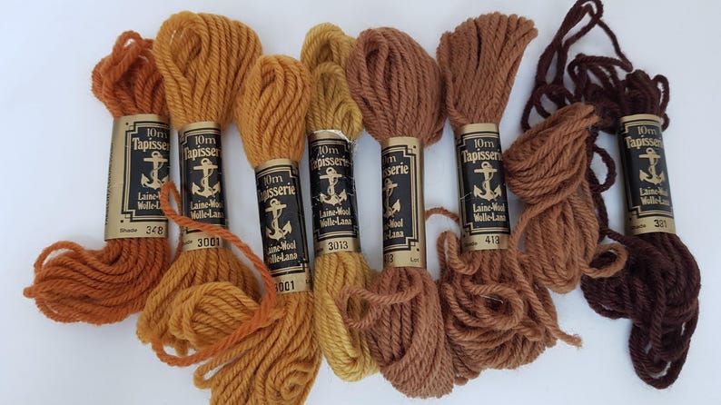 Pode incluir: Seis novelos de l&atilde; para tape&ccedil;aria de 10 mm em v&aacute;rios tons de marrom e laranja. A l&atilde; &eacute; etiquetada como "10mm Tapisserie Laine-Wool Wolle-Lana" com um n&uacute;mero em cada novelo.