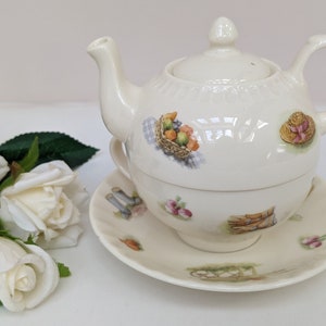 May include: A white ceramic teapot and cup set with a floral design. The teapot has a lid and a handle. The cup has a saucer. The set features a floral design with a basket of fruit, a hat, and other details.