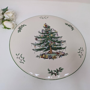 May include: A white ceramic cake stand with a green rim and a Christmas tree design. The tree is decorated with baubles and presents.