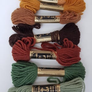 Pode incluir: Dez novelos de l&atilde; Anchor Tapisserie em v&aacute;rias cores, incluindo branco, amarelo, laranja, castanho, vermelho, verde e preto. Cada novelo &eacute; etiquetado com o logotipo da Anchor, "Tapisserie", "Wool-Laine" e o n&uacute;mero da cor.