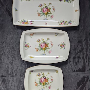 May include: Three white ceramic serving trays with gold trim and floral designs. The trays are different sizes and feature a variety of colourful flowers.