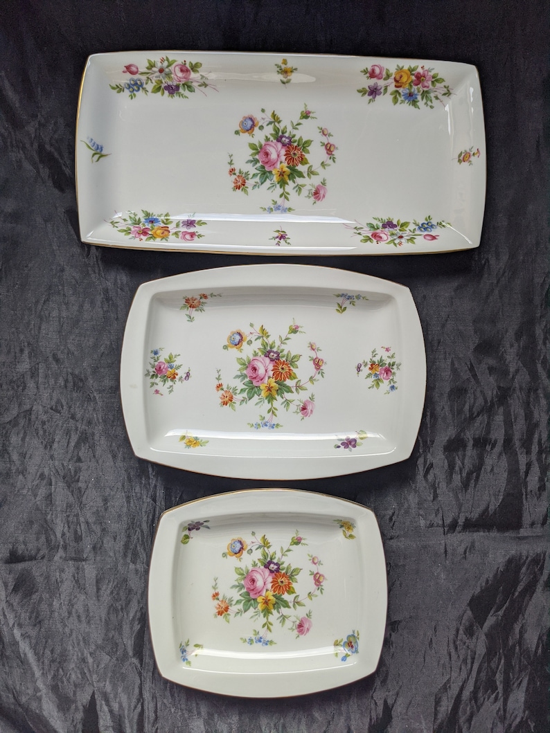 May include: Three white ceramic serving trays with gold trim and floral designs. The trays are different sizes and feature a variety of colourful flowers.