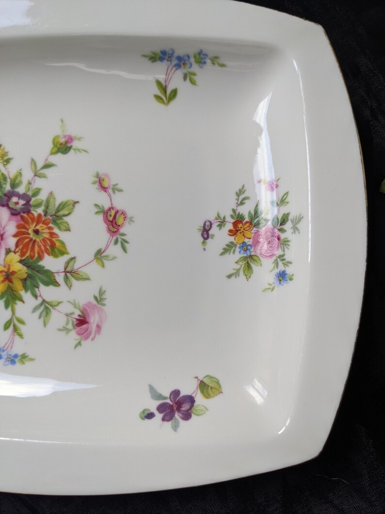 May include: White ceramic serving dish with a square shape and rounded corners. The dish is decorated with a floral pattern in shades of pink, yellow, orange, blue, and purple.