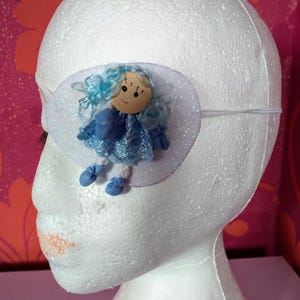 May include: A white eye patch with a blue and white doll applique. The doll has blue hair, a blue dress, and blue shoes.
