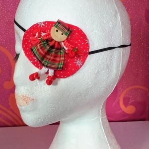 May include: A red eye patch with a white and green tartan doll applique. The doll has a red hat and a wooden face. The eye patch is attached to a black elastic band.