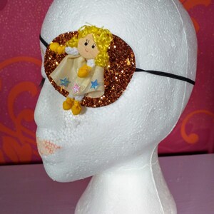 May include: A gold glitter eye patch with a felt angel doll applique. The doll has blonde hair, a white dress, and yellow shoes.