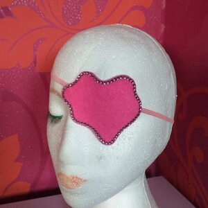 May include: A pink heart-shaped eye patch with rhinestones around the edge. The patch is attached to an elastic headband.
