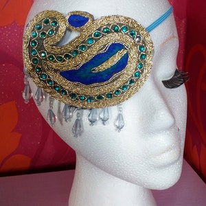 May include: A white mannequin head with a blue and gold eye patch with dangling crystal beads. The eye patch is decorated with blue and green jewels.