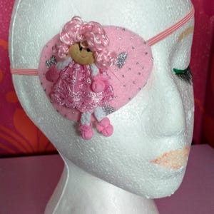 May include: A pink eye patch with a felt doll applique. The doll has pink hair, a pink dress, and white shoes. The eye patch is attached to a pink elastic band.