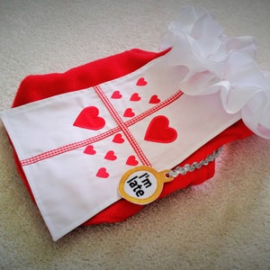 May include: A red and white garment featuring a white ruffled collar and a white panel decorated with red hearts and a red checkered cross. A circular yellow tag with a silver chain is attached, displaying the black text I'm late.