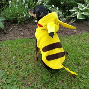 May include: A bright yellow fleece pet costume featuring a hood with long pointed ears, black ear tips, and red circular cheek patches. The back of the garment displays two horizontal dark brown stripes and a long yellow tail with a lightning bolt shape.