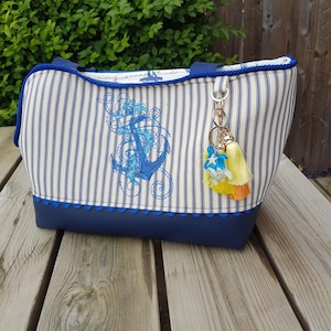 May include: A blue and white striped tote bag with a nautical anchor design. The bag has a blue scalloped trim, a blue handle, and a decorative tassel keychain. The bag is made of fabric and has a rectangular shape.