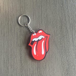 May include: A red and white Rolling Stones tongue and lips logo keyring. The keyring features a silver metal ring and chain. The tongue and lips design is a classic rock and roll symbol.