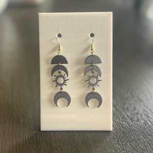 Puede incluir: Pendientes grises oscuros con un diseño celestial. Cada pendiente presenta un semicírculo, una luna creciente y un colgante de sol, conectados por ganchos dorados. Los pendientes se exhiben en un soporte rectangular de color crema.