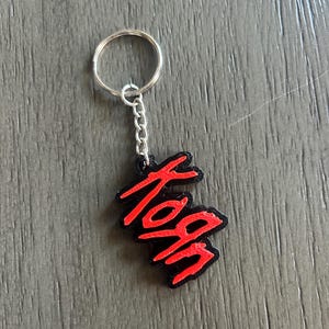 Puede incluir: Un llavero con una base negra y la palabra Korn en relieve en letras de color rojo brillante. El diseño está unido a una anilla de metal plateado mediante una cadena corta, creando un accesorio distintivo con un fuerte contraste.