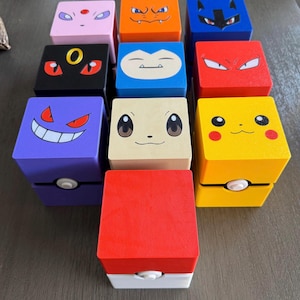 Puede incluir: Una colección de diez cajas de almacenamiento cuadradas y multicolores con varios rostros de personajes. Cada caja muestra un diseño único en colores amarillo, rojo, azul, morado, negro, naranja y rosa, con algunas cajas que incluyen un pequeño botón blanco.