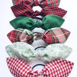 May include: A collection of six fabric hair ties with bow accents. The bows feature various patterns, including red and green plaid, green solid, white with green fern-like prints, and red gingham. Each bow is attached to a black elastic band.