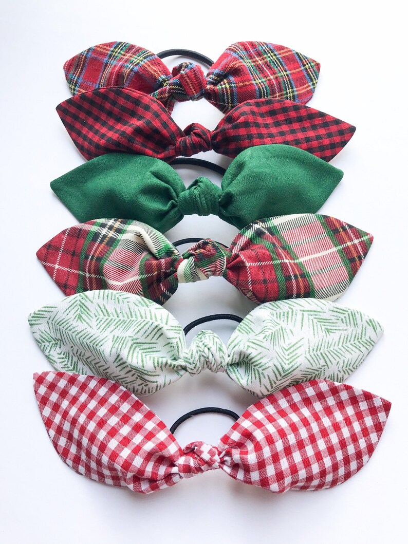 May include: A collection of six fabric hair ties with bow accents. The bows feature various patterns, including red and green plaid, green solid, white with green fern-like prints, and red gingham. Each bow is attached to a black elastic band.