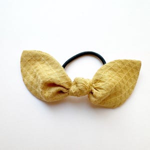 May include: Mustard yellow fabric hair tie with a textured waffle weave pattern. The fabric is tied into a bow shape, attached to a black elastic band. The hair accessory is set against a white background.