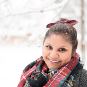 May include: A smiling person wearing a red, green, and black plaid scarf and a matching bow in their hair. The person is wearing a gray sweater. The background is a snowy outdoor scene.