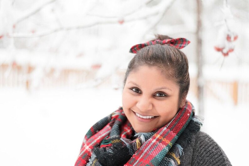 May include: A smiling person wearing a red, green, and black plaid scarf and a matching bow in their hair. The person is wearing a gray sweater. The background is a snowy outdoor scene.