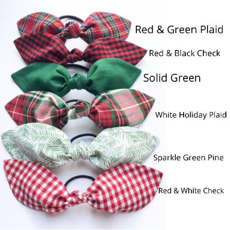 May include: A collection of six hair ties with bow accents. The bows feature various patterns, including red and green plaid, red and black check, solid green, white holiday plaid, sparkle green pine, and red and white check.