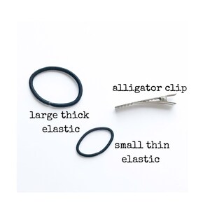 May include: An assortment of hair accessories on a white background. The image includes a large, thick black elastic hair tie, a small, thin black elastic hair tie, and a silver alligator clip. Text labels identify each item.