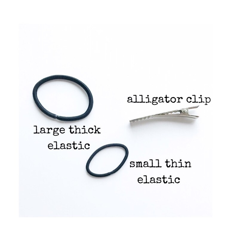 May include: An assortment of hair accessories on a white background. The image includes a large, thick black elastic hair tie, a small, thin black elastic hair tie, and a silver alligator clip. Text labels identify each item.