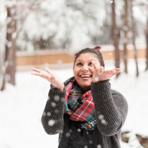 May include: A person wearing a gray sweater and a red, green, and black plaid scarf, smiling and looking up at falling snow. The background shows snow-covered trees and a wooden fence.
