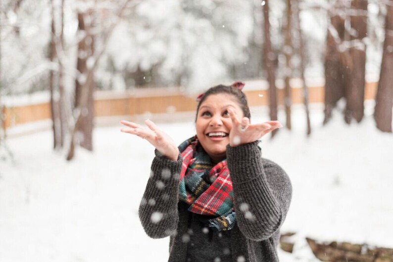 May include: A person wearing a gray sweater and a red, green, and black plaid scarf, smiling and looking up at falling snow. The background shows snow-covered trees and a wooden fence.