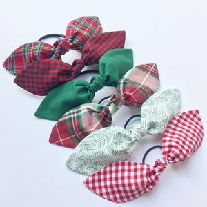 May include: A collection of six hair ties with fabric bows in various patterns and colors. The bows include red plaid, green, and white with red and green patterns. Each bow is attached to a black elastic band.