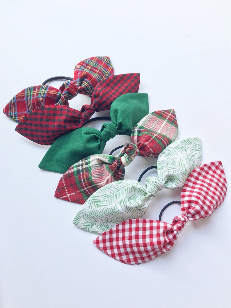 May include: A collection of six hair ties with fabric bows in various patterns and colors. The bows include red plaid, green, and white with red and green patterns. Each bow is attached to a black elastic band.