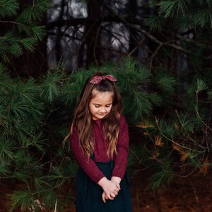 May include: A young person with long brown hair wearing a burgundy long-sleeve shirt and a dark green skirt. A red plaid bow is in their hair. The background features green pine branches and a dark forest.