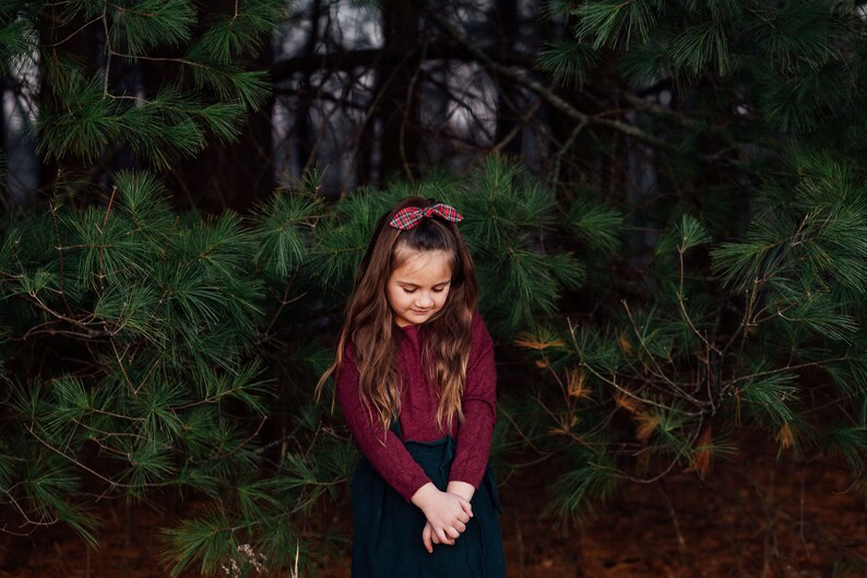 May include: A young person with long brown hair wearing a burgundy long-sleeve shirt and a dark green skirt. A red plaid bow is in their hair. The background features green pine branches and a dark forest.