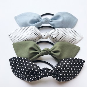 May include: Four fabric hair scrunchies in a stack. The top scrunchie is light blue, the second is white with black polka dots, the third is olive green, and the bottom is black with white polka dots.