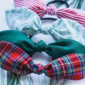 May include: A collection of fabric hair ties in various patterns and colors. The assortment includes red and white striped, green leaf print, solid green, and red plaid designs. The hair ties are arranged on a white surface.