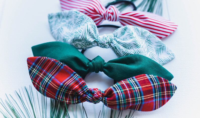 May include: A collection of fabric hair ties in various patterns and colors. The assortment includes red and white striped, green leaf print, solid green, and red plaid designs. The hair ties are arranged on a white surface.