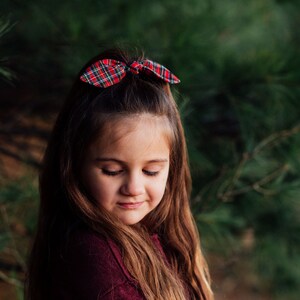 May include: A young person with long brown hair and a red and green plaid bow in their hair. The bow is tied in a bunny ear style. The person is wearing a burgundy top, and the background is a blurred green.
