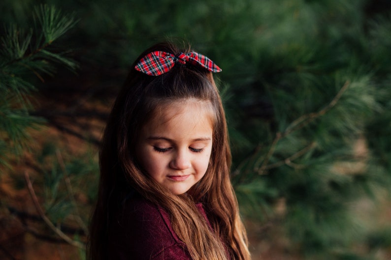 May include: A young person with long brown hair and a red and green plaid bow in their hair. The bow is tied in a bunny ear style. The person is wearing a burgundy top, and the background is a blurred green.